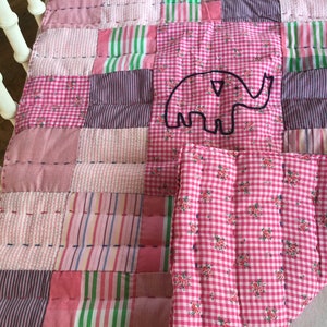 Peut inclure: Une couverture patchwork pour bébé rose, composée de carrés de différents tissus, dont du vichy et des rayures. Un éléphant brodé est centré sur un carré vichy. La couverture mesure probablement 100 x 127 cm.