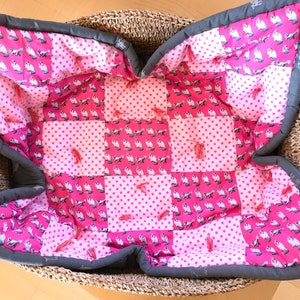 Puede incluir: Una cama acolchada para mascotas de color rosa y blanco con un borde gris. La cama está hecha de tela de patchwork con un patrón de lunares y un patrón de pequeños animales blancos sobre un fondo rosa.