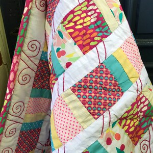 Peut inclure: Une courtepointe colorée en patchwork avec un motif en losange. La courtepointe est faite de différents tissus avec des motifs différents, y compris des pois, des rayures et des motifs floraux. La courtepointe est pliée en deux et est accrochée à une porte.