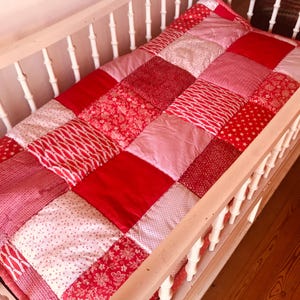 Op de afbeelding: Een rood en roze patchwork quilt in een wieg. De quilt is gemaakt van verschillende stoffen, waaronder effen kleuren, bloemenprints en stippenpatronen. De quilt is rechthoekig en lijkt handgemaakt.