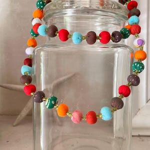 May include: A vibrant necklace featuring fabric-covered beads in shades of red, orange, green, and blue. The beads are strung on a gold-coloured string. The necklace is displayed over a clear glass jar.