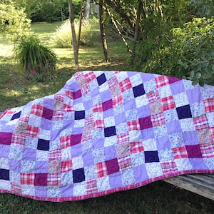 Può includere: Una trapunta patchwork con quadrati di vari motivi floreali e a quadri in tonalità di rosa, viola e bianco. La trapunta è realizzata in tessuto di cotone ed è appoggiata su una panca di legno.