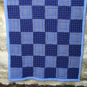 Op de afbeelding: Een blauw en wit geruit patchworkdeken met een lichtblauwe rand. Het deken is gemaakt van vierkanten met verschillende geruite patronen.