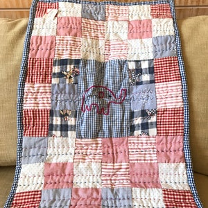 May include: A patchwork baby quilt with a red, white, and blue color scheme. The quilt is made of squares of different fabrics, including gingham, stripes, and polka dots. An embroidered elephant is in the center of the quilt.
