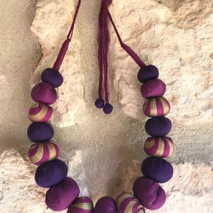 May include: A necklace featuring a series of round, fabric-covered beads in solid deep purple and alternating purple and gold striped patterns. The beads are strung on a thin, dark purple cord that includes a small wooden bead at the top closure.