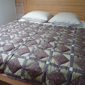 Peut inclure: Une couette matelassée marron et verte avec un motif en losange. La couette est sur un lit avec deux oreillers blancs.