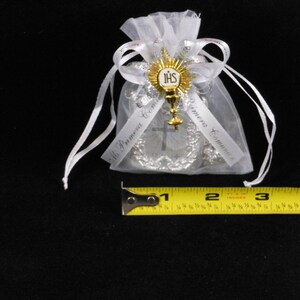 May include: White organza bag with a silver and gold religious charm. The charm features a cross, a chalice, and the letters "JHS". The bag is tied with a white ribbon and has the text "Mi Primera Comuni&oacute;n" printed on it.