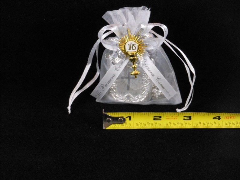 May include: White organza bag with a silver and gold religious charm. The charm features a cross, a chalice, and the letters "JHS". The bag is tied with a white ribbon and has the text "Mi Primera Comuni&oacute;n" printed on it.