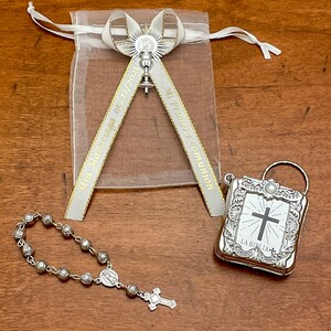 May include: A silver rosary with a small silver cross charm, a silver book with a cross and the text "LA BIBLIA" on the cover, and a white organza bag with a silver charm and a ribbon that says "My First Communion" and "May 02, 2021".