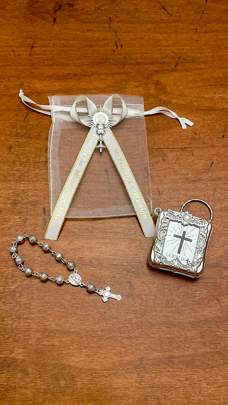 May include: A silver rosary with a small silver cross charm, a silver book with a cross and the text "LA BIBLIA" on the cover, and a white organza bag with a silver charm and a ribbon that says "My First Communion" and "May 02, 2021".