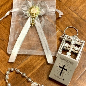 May include: A silver rosary with a small silver charm, a silver keychain with a small book that says "Dios Habla Hoy La Biblia" and a white organza bag with a silver ribbon and a gold charm.