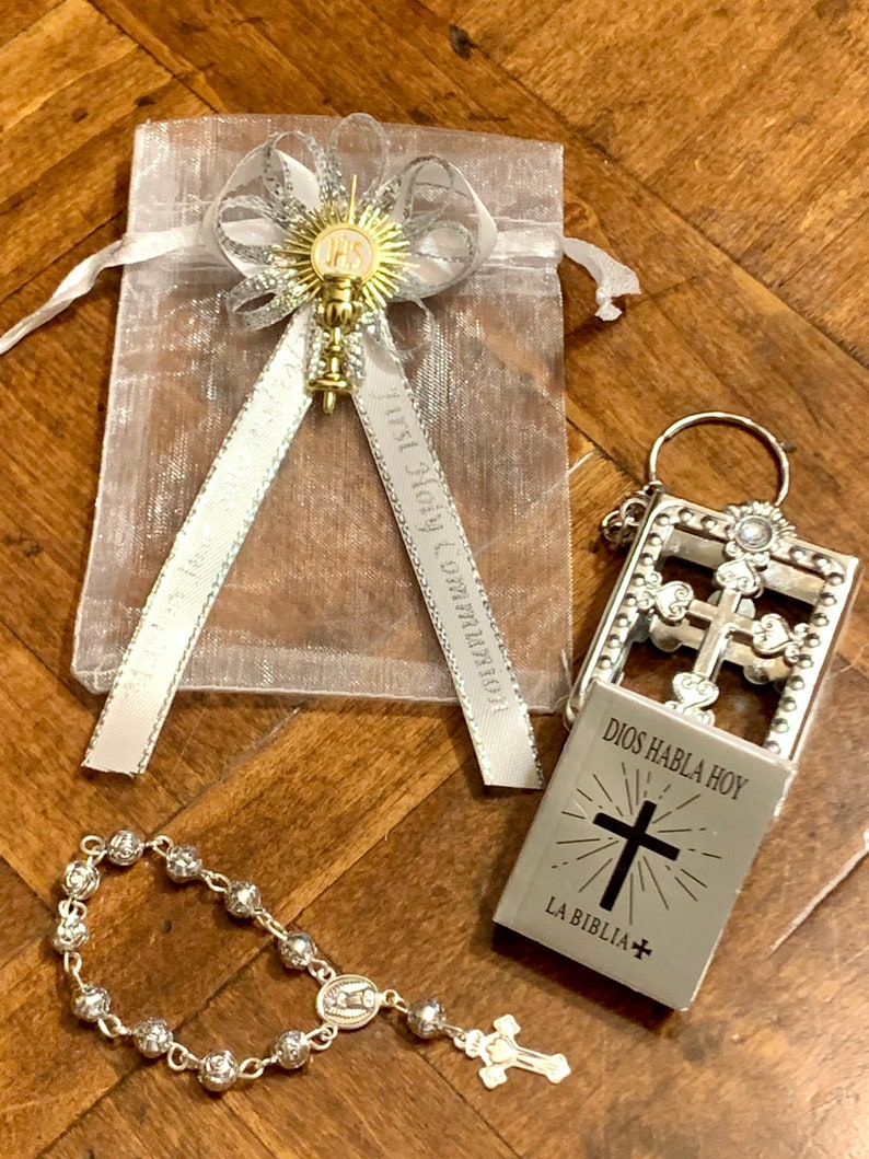 May include: A silver rosary with a small silver charm, a silver keychain with a small book that says "Dios Habla Hoy La Biblia" and a white organza bag with a silver ribbon and a gold charm.