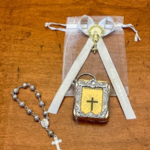 Könnte beinhalten: Eine silberne Rosenkranz mit einem silbernen Kreuzanhänger, eine silberne Metall-Bibel mit einem Kreuz auf der Vorderseite und eine weiße Organza-Tasche mit einem silbernen Band und einem goldenen Kreuzanhänger. Das Band trägt den Text "Erste Heilige Kommunion" aufgedruckt.