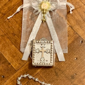 May include: A silver and gold religious gift set for a first communion. The set includes a small silver book with a cross and the words "Dios te ama hoy" on the front, a silver rosary with a silver cross and a silver charm, and a silver and gold charm with a chalice and the letters "IHS" on a white ribbon.