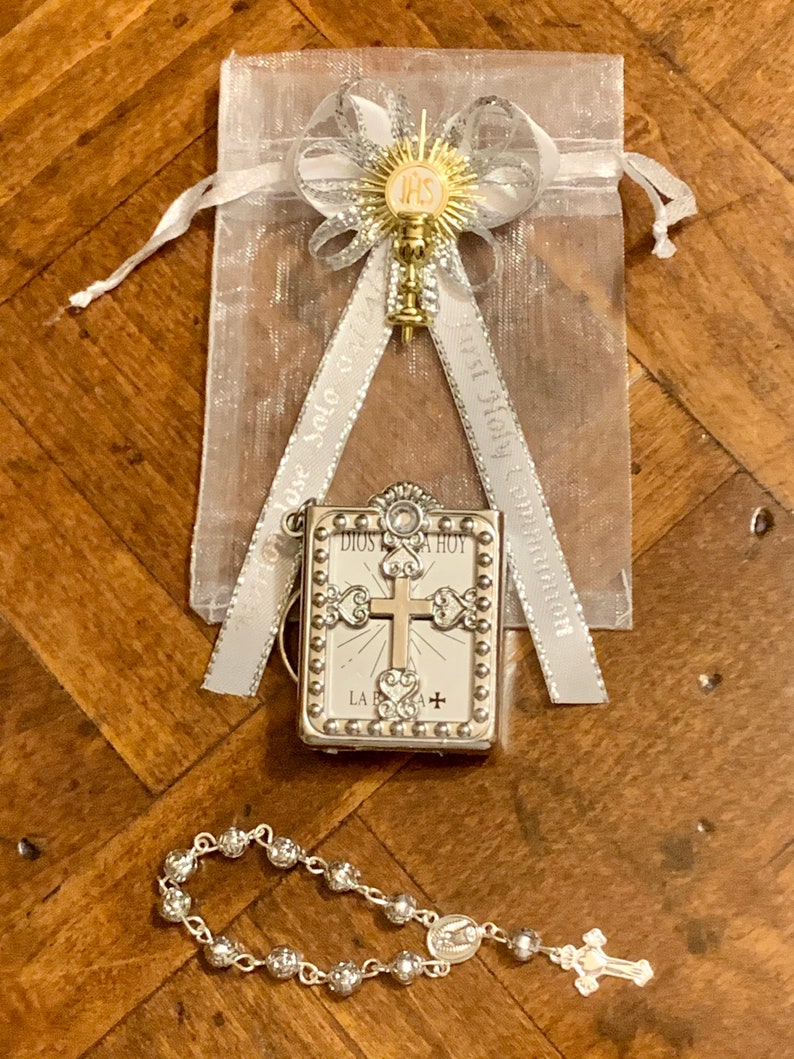 May include: A silver and gold religious gift set for a first communion. The set includes a small silver book with a cross and the words "Dios te ama hoy" on the front, a silver rosary with a silver cross and a silver charm, and a silver and gold charm with a chalice and the letters "IHS" on a white ribbon.