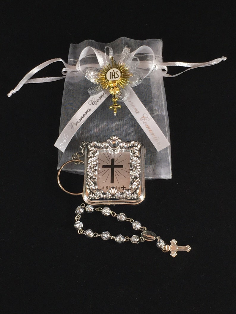 May include: A silver-colored book charm with a cross and the words "La Biblia" on the front, a silver rosary with a cross charm, and a white organza bag with a gold and silver ribbon that says "Mi Primera Comuni&oacute;n" on it.