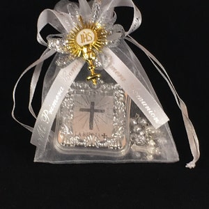 May include: A white organza bag with a silver ribbon and a silver and gold religious charm. The charm features a cross and the words "La Biblia" and "IHS".