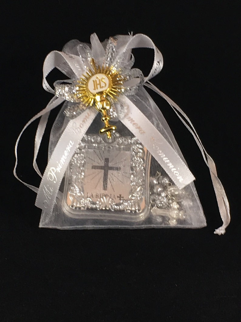 May include: A white organza bag with a silver ribbon and a silver and gold religious charm. The charm features a cross and the words "La Biblia" and "IHS".