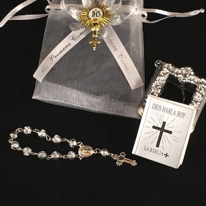 May include: A white organza bag with a gold and silver ribbon that says "Mi Primera Comuni&oacute;n". The bag contains a small silver book with the words "Dios Habla Hoy" and "La Biblia" on the cover, and a silver rosary with a small cross.
