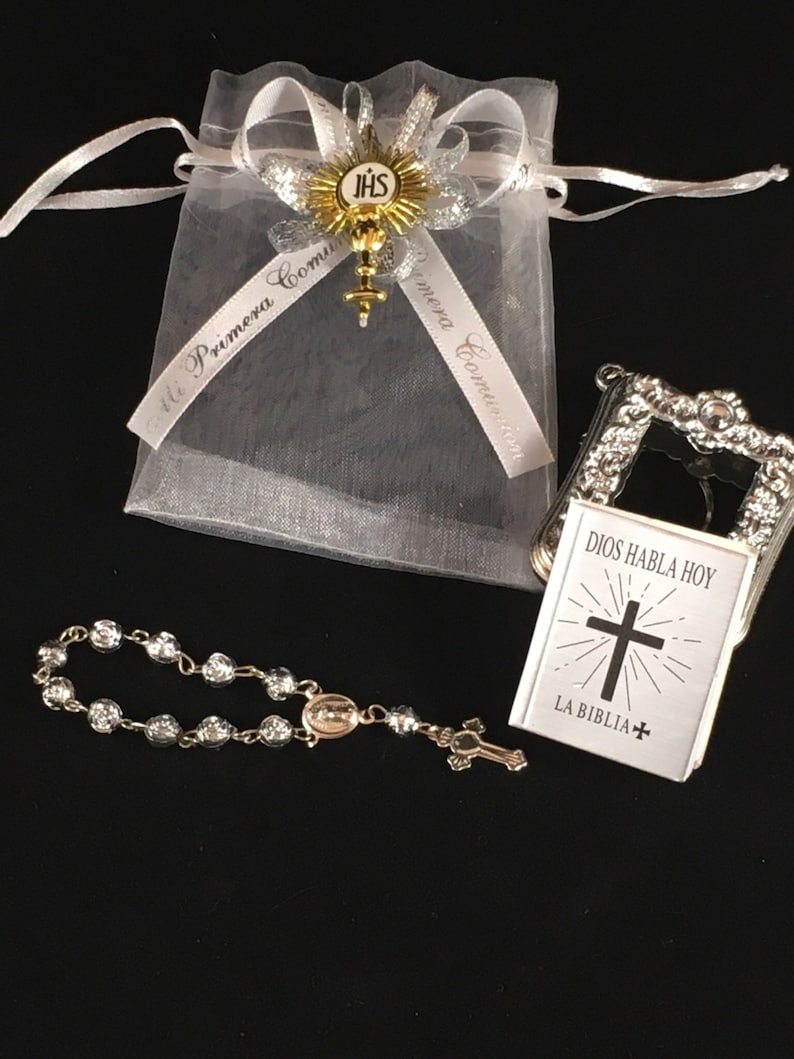 May include: A white organza bag with a gold and silver ribbon that says "Mi Primera Comuni&oacute;n". The bag contains a small silver book with the words "Dios Habla Hoy" and "La Biblia" on the cover, and a silver rosary with a small cross.