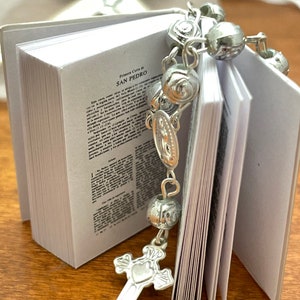 May include: A small white book with the text "Primera Carta de SAN PEDRO" on the cover. A silver rosary with a silver cross charm is draped over the book.