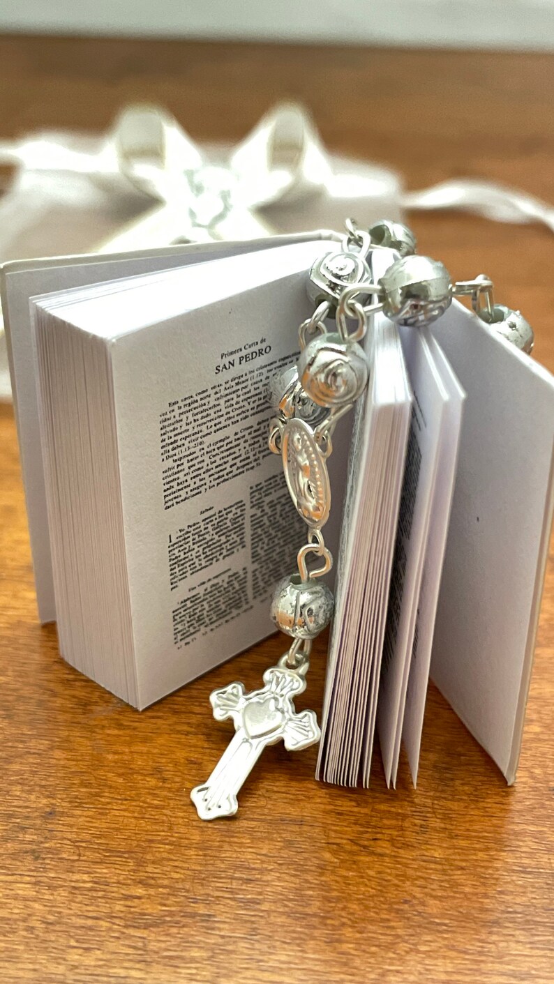May include: A small white book with the text "Primera Carta de SAN PEDRO" on the cover. A silver rosary with a silver cross charm is draped over the book.