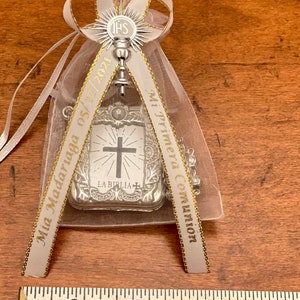 May include: A white satin bag with gold trim and a silver charm. The charm is a cross with a bible verse and the words "Mi Primera Comunion" and "Mia Madariaga 05/05" are printed on the ribbon.