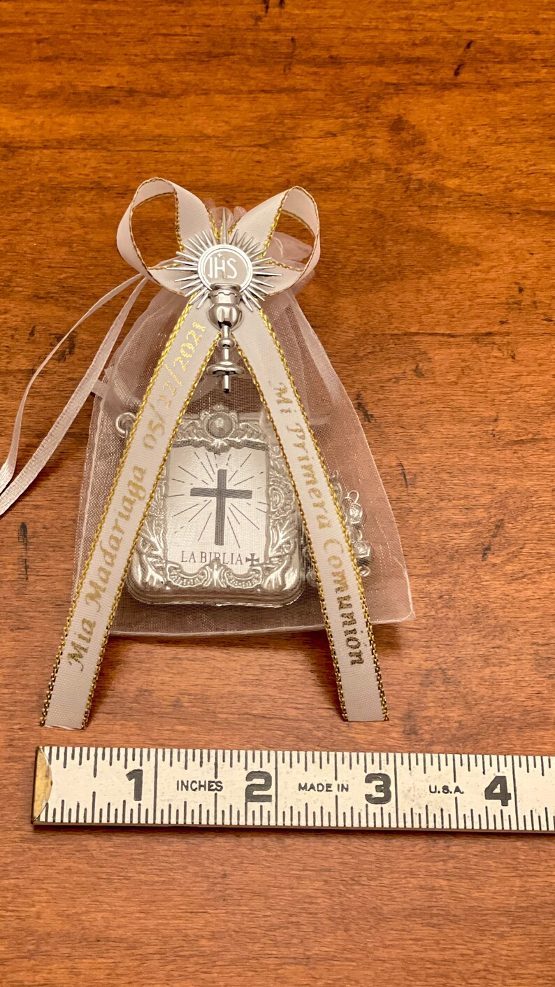 May include: A white satin bag with gold trim and a silver charm. The charm is a cross with a bible verse and the words "Mi Primera Comunion" and "Mia Madariaga 05/05" are printed on the ribbon.