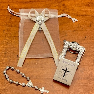 May include: A silver charm with a cross and the text "Dios Habla Hoy" and "La Biblia" on a white background. The charm is attached to a silver key chain. A silver rosary with a silver cross and a silver charm with the Virgin Mary is also pictured. A white organza bag with a silver charm and a gold ribbon with the text "Mi Primera Comuni&oacute;n" and "05.12.2023" is also pictured.