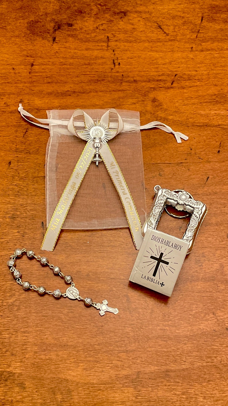 May include: A silver charm with a cross and the text "Dios Habla Hoy" and "La Biblia" on a white background. The charm is attached to a silver key chain. A silver rosary with a silver cross and a silver charm with the Virgin Mary is also pictured. A white organza bag with a silver charm and a gold ribbon with the text "Mi Primera Comuni&oacute;n" and "05.12.2023" is also pictured.