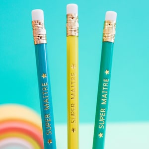 Peut inclure: Trois crayons de couleur bleu, jaune et vert, chacun avec une gomme blanche et une virole en métal doré. Les crayons portent l'inscription "SUPER MAITRE" et une étoile.