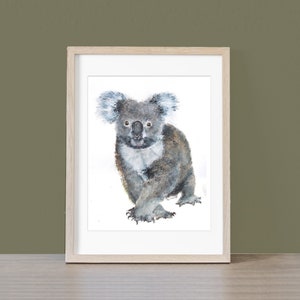 May include: Watercolor painting of a koala bear in a wooden frame. The koala is depicted in shades of gray and brown, with fluffy ears and a focused expression. The artwork is set against a white background.