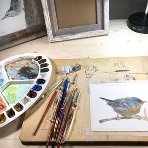 May include: A watercolor painting of a bluebird perched on a branch, with a palette of watercolors, brushes, and a tube of paint on a wooden table.