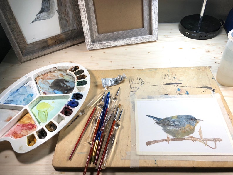 May include: A watercolor painting of a bluebird perched on a branch, with a palette of watercolors, brushes, and a tube of paint on a wooden table.