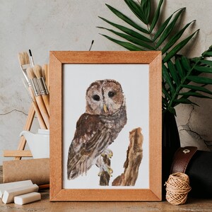 May include: A framed watercolor painting of a brown owl perched on a branch. The painting is in a wooden frame and is sitting on a wooden table with art supplies.