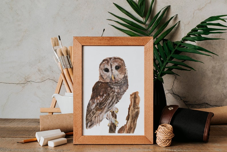 May include: A framed watercolor painting of a brown owl perched on a branch. The painting is in a wooden frame and is sitting on a wooden table with art supplies.