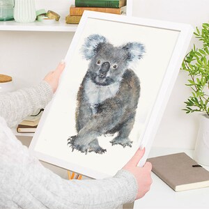 May include: A framed watercolor print of a koala bear. The koala is depicted in shades of gray and white, with a fluffy appearance. The artwork is set against a white background and framed in a white wooden frame.