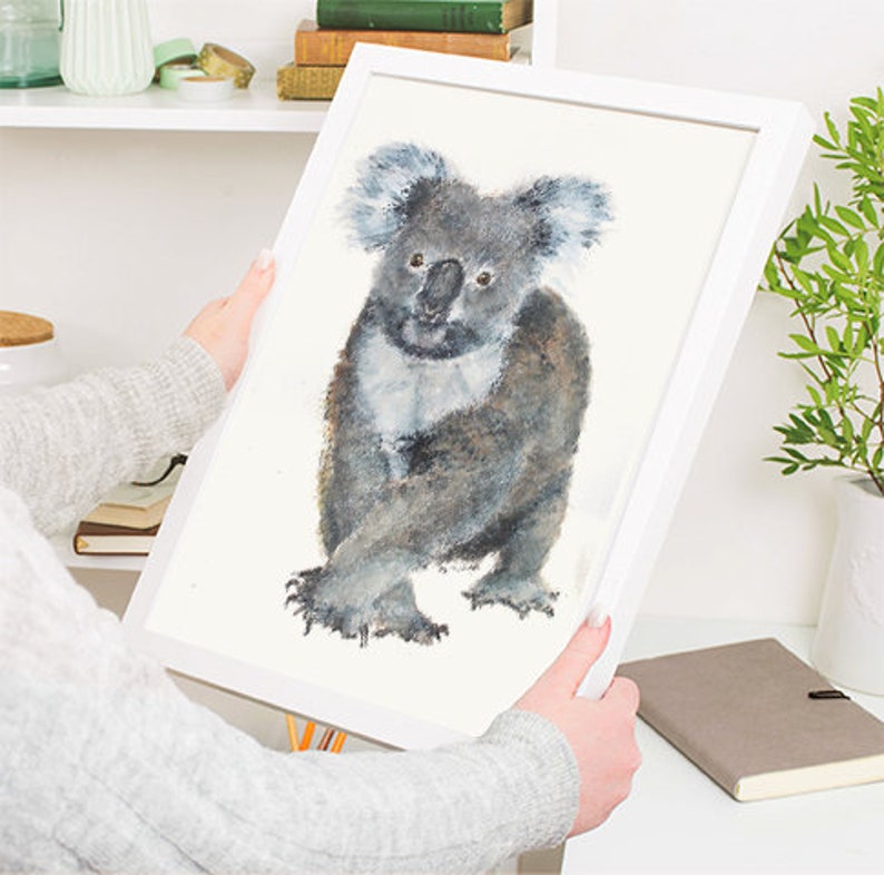 May include: A framed watercolor print of a koala bear. The koala is depicted in shades of gray and white, with a fluffy appearance. The artwork is set against a white background and framed in a white wooden frame.