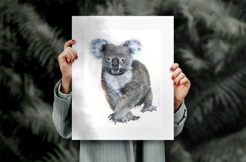 May include: Watercolor painting of a koala bear. The koala is depicted in shades of gray and blue, with a fluffy appearance. The artwork is held by a person with red nail polish, against a blurred, dark green background.