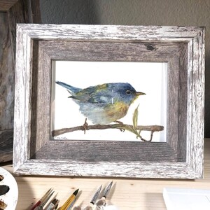May include: A watercolor painting of a blue, yellow, and green bird perched on a branch. The painting is framed in a rustic wooden frame with a whitewashed finish.