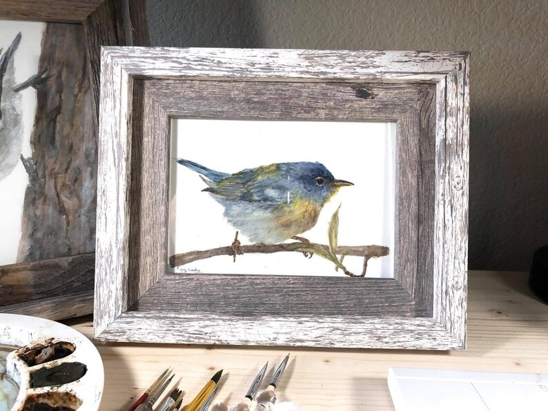 May include: A watercolor painting of a blue, yellow, and green bird perched on a branch. The painting is framed in a rustic wooden frame with a whitewashed finish.