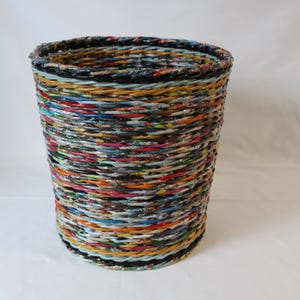 May include: A cylindrical waste paper basket constructed from tightly woven, multi-coloured paper strips. The basket displays a variety of colours such as red, blue, yellow, and green, giving it a vibrant and textured look. The top and bottom edges are reinforced with a darker material.