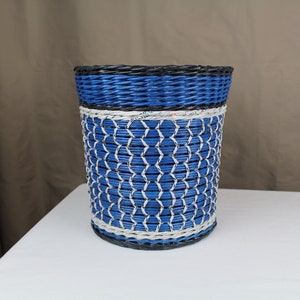 May include: A blue and black woven wastebasket with a white stripe. The basket is made of paper and has a woven pattern.