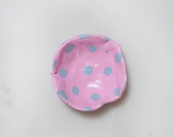 May include: A small pink ceramic dish with a polka dot pattern in light blue. The dish is slightly misshapen, giving it a handmade look.
