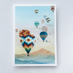 May include: Watercolor painting of hot air balloons in flight. The balloons are various colors and patterns, including rainbow, blue, and striped designs. The background features a light blue sky, mountains, and a soft, watercolor effect.