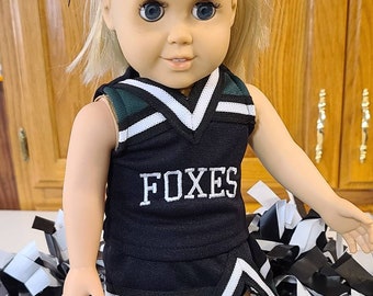 May include: A doll wearing a black and green cheerleader uniform with the word "FOXES" in white. The doll has blonde hair and a black bow. Black and white pom-poms are in the background.