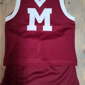 May include: Maroon cheerleading uniform set with white trim. The top has a white "M" and a V-neck design. The skirt is a matching maroon color with a white border. The uniform is made of a smooth, shiny fabric.