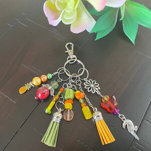 May include: A colourful keychain with a silver key ring and multiple charms, including a pineapple, a flower, a butterfly, and a tassel. The charms are made of glass, metal, and fabric.