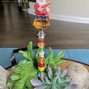 K&ouml;nnte beinhalten: Eine fantasievolle Gartenzwergfigur, die ein Willkommensschild h&auml;lt, thront auf einem kristallartigen Aufsatz mit einem Perlenstiel. Der Zwerg tr&auml;gt ein rotes und blaues Outfit und ist von gr&uuml;nen Sukkulenten umgeben.
