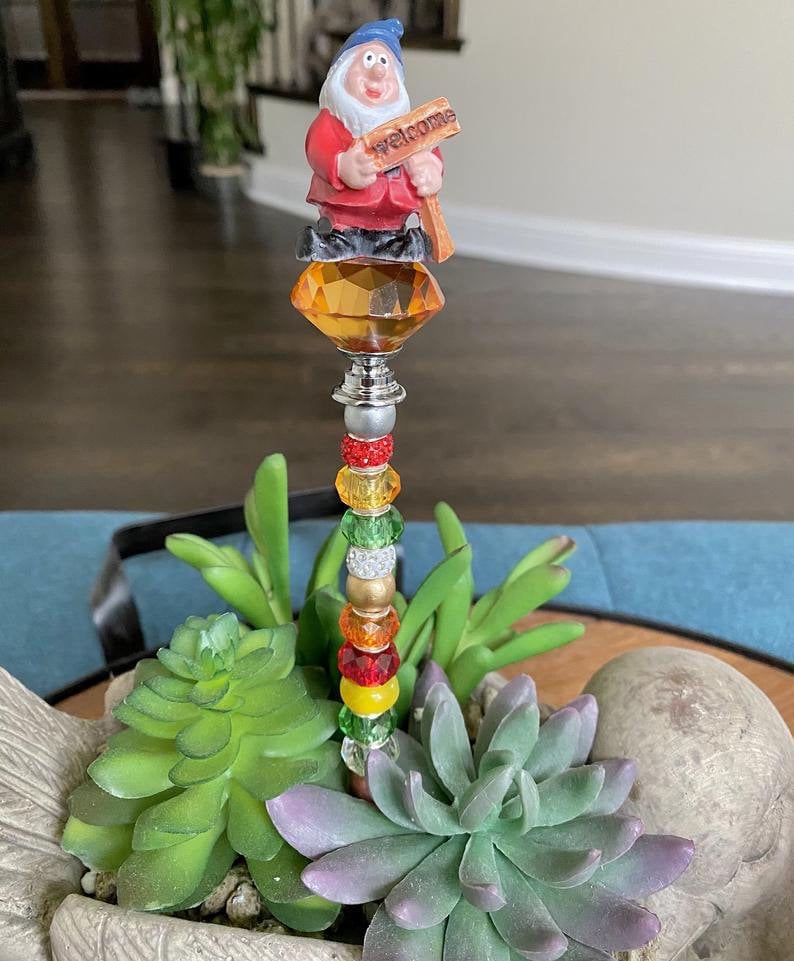 K&ouml;nnte beinhalten: Eine fantasievolle Gartenzwergfigur, die ein Willkommensschild h&auml;lt, thront auf einem kristallartigen Aufsatz mit einem Perlenstiel. Der Zwerg tr&auml;gt ein rotes und blaues Outfit und ist von gr&uuml;nen Sukkulenten umgeben.